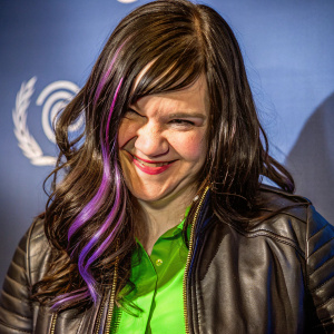 Caucasian actress Lena Hall, cheerful and vibrant, wearing a green shirt and black leather jacket. Purple streaks in her long wavy dark hair. Against a light-colored backdrop with subtle logo-like design. Soft, even lighting highlights her features. Close-up perspective focusing on her upper body and face.