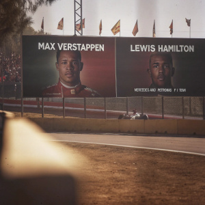 Max Verstappen and Lewis Hamilton's giant billboards at Formula 1 race. Crowded venue, sunny day.
