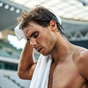 Rafael Nadal, shirtless, intense focus. Wiping sweat with a white towel. Indoor stadium background, blurred