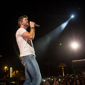 Robin Thicke, wearing a white t-shirt and jeans, performing under bright spotlights at Coachella. The crowd is cheering.