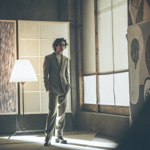 Timothée Chalamet, wearing a green suit with subtle shimmer, standing in a dimly lit studio with abstract art pieces in the background.