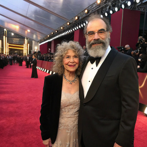 Mandy Patinkin and Kathryn Grody at the award ceremony, standing on the vibrant red carpet. Mandy, with his iconic beard, and Kathryn, with her charming curly hair, are posing for photographers.