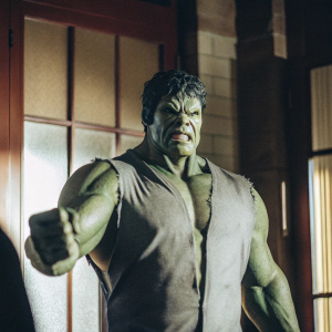 Green-skinned Marvel hero Hulk, standing with a powerful stance. His muscular body is well-defined, with a broad chest and wide shoulders. Wearing a torn gray shirt. Standing in a dimly lit room with a warm brown background.