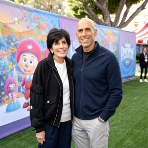 Marilyn Katzenberg, in a casual outfit, and Jeffrey Katzenberg posing at an outdoor event. They stand in front of a colorful banner featuring DreamWorks characters.