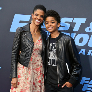 Renée Elise Goldsberry and her son Benjamin Johnson at the 'Fast and Furious: Spy Racers' premiere. Renée in a floral-patterned dress with quilted design and leather jacket. Benjamin in graphic t-shirt and leather jacket. Both smiling. Dark backdrop with graphic elements, high-resolution photo
