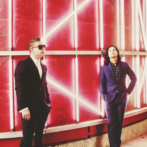 Skrillex and Diplo, standing at the Billboard Music Awards, both dressed in formal attire. Skrillex in black suit with sunglasses, Diplo in dark blue suit. They pose in front of a neon-lit backdrop featuring the Billboard logo.