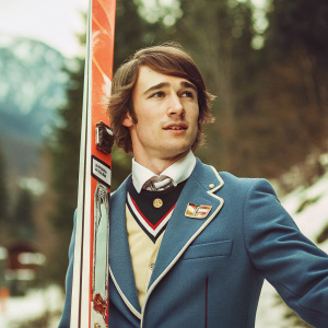 Kamil Stoch, Polish ski jumper, at the Winter Olympics. Wearing blue suit with sponsor logos, posing with a Fischer ski. Snowy landscape backdrop