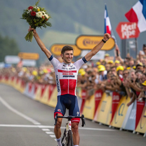 Giulio Ciccone, standing victorious at the finish line of a grand cycling event. Celebratory pose with bouquet and sponsor banner. Crowd cheering in background. High-definition photo.
