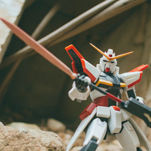 An action figure of Gundam Astray Red Frame, striking a powerful pose with a weapon in hand, set against a black backdrop.