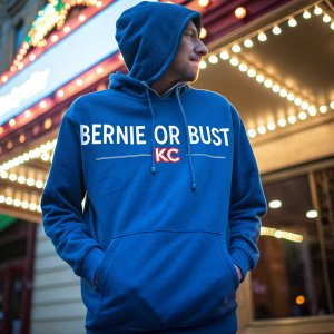 A blue hoodie with Bernie Sanders campaign slogan '#BernieorBustKC' and Kansas City city emblem. Brightly lit background