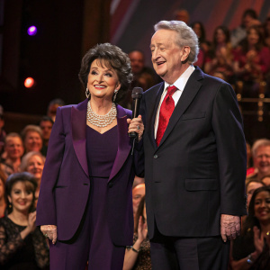 Suzanne Pleshette and Tom Poston at a prestigious charity gala. They both stand on stage, Suzanne in a dark purple blazer and pearl necklace, Tom in a dark suit with a red tie. Crowd cheering