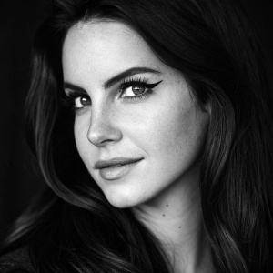 Close-up black and white photo of Lana Del Rey. Smirking with intense gaze. Defined eyebrows, long dark hair, and smooth skin. Dark background highlights her features