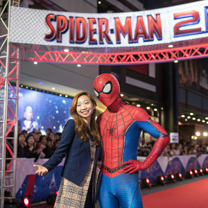 Tomomi Itano, a Japanese idol, posing with Spider-Man mascot. Event stage with audience visible. Red and blue decorations. Promotional banner for 'Amazing Spider-Man 2' on the backdrop.