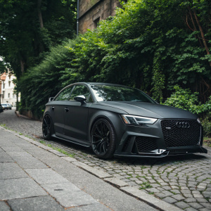 High-performance Audi RS3 car in matte black parked on a cobblestone street surrounded by lush greenery. The distinctive grille pattern and black accents are prominently visible.