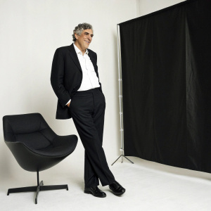 Elliott Gould, dressed in a classic black suit and crisp white shirt, strikes a friendly pose in a minimalist studio setting with a sleek black backdrop.