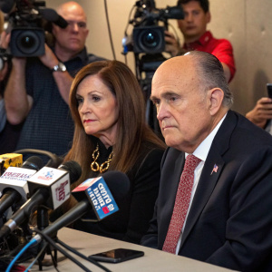 Rudy Giuliani and Judith Giuliani at a press conference. Sitting at a table with microphones in front of them. News cameras and reporters in the background.