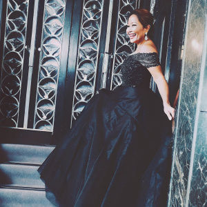 Carrie Ann Inaba, smiling brightly at the Met Gala. Elegant black gown with delicate lace. Background features intricate gold designs.