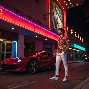Mariano Di Vaio standing beside a sleek red Ferrari. Nightclub exterior in neon lights. Wearing a patterned shirt and white pants.