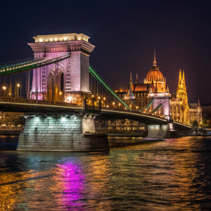 At night, Szechenyi Chain Bridge in Budapest lights up in a dazzling array of colors. Ornate architectural details of the bridge and its reflection in the Danube River create a stunning visual spectacle.