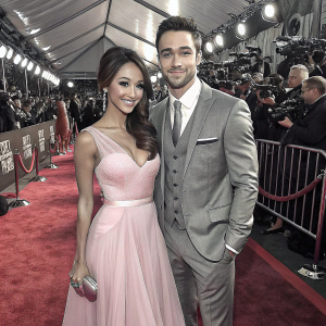 Melanie Iglesias, wearing a pastel pink, sleeveless gown with a sheer overlay, and Ryan Guzman, in a gray suit, attend the People's Choice Awards. Red carpet scene with flashing cameras and paparazzi.