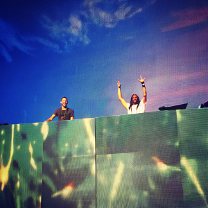 Nicky Romero and Steve Aoki at the EDM concert. Wearing casual stage outfits. Digital backdrop with vibrant graphics.