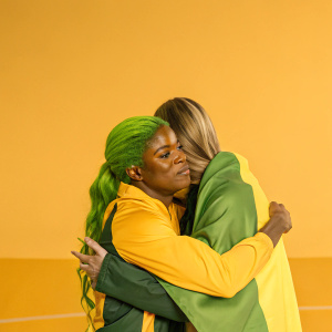 Shelly-Ann Fraser-Pryce and Dafne Schippers in a heartfelt embrace. Dimly lit indoor stadium, both athletes wearing sportswear. Fraser-Pryce has vibrant green hair. Schippers looks worried.