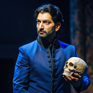A man. neatly trimmed black hair.  Blue. Hamlet's pose. Skull. Background: Theatre stage