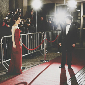 Sandra Oh and Alexander Payne at a film festival premiere. Both dressed in formal attire. Red carpet event with flashing cameras and crowds.