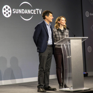 David Remnick and Esther Fein attending the SundanceTV press conference. The stage is set against a sleek gray backdrop with event logos and text