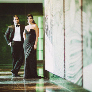Henry Cavill and Gina Carano at a gala. He in tux, she in black gown. They stand confidently against backdrop of abstract art. Dimly lit room with glossy floors.