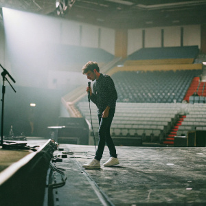 Nathan Sykes performing on a stage. Male singer with short brown hair. Dark stage lighting. Crowd not visible