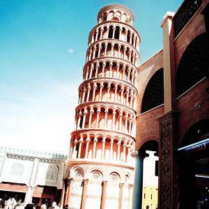 Replica of the Leaning Tower of Pisa at Global Village Dubai. Rich, golden-brown hue, leaning slightly. Multi-level design with arches and spiral pattern. Historic Italian style. Bustling area with people and shops in the background. Clear, bright blue sky.