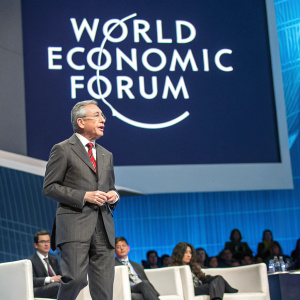 Alvaro Uribe Velez, ex-President of Colombia, delivering a keynote speech at the World Economic Forum. Standing confidently on stage, wearing a dark grey suit with a red tie. Behind him is a large, modern digital screen displaying the forum's logo.