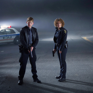 Jennifer Lopez and Warren Kole in intense scene from 'Shades of Blue'. Dimly lit street with police car in background. Both actors are in uniform, holding guns, with serious expressions.