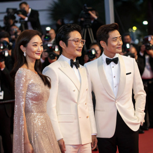 Han Chae-Young, Lee Jae-Gyu, and Lee Byung-Hun at the Grand Ballroom premiere. Chae-Young in shimmering gown, Jae-Gyu in white suit, Byung-Hun in classic tuxedo. Paparazzi flashing lights in the background.