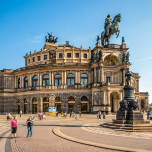 Semperoper, the grand opera house in Dresden, featuring elaborate architecture with multiple arches and a central statue of a figure on horseback. Lively scene on a sunny day, filled with tourists and locals.