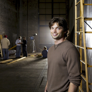 Tom Welling, casual in a brown sweater, on set between takes. The bustling film studio background, filled with crew members and equipment.