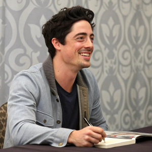 Ben Feldman, actor, at a book signing event. Cheerful demeanor, casual outfit. Gray-patterned backdrop.