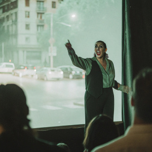 Caucasian woman, Rossy de Palma, dramatically posing on a dimly lit stage. The backdrop features a bustling cityscape at night