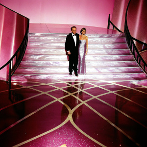Rupert Murdoch and Juliet de Baubigny posing on a luxurious platform with intricate floor design at the Vanity Fair Oscar Party. The backdrop is adorned with shimmering details.