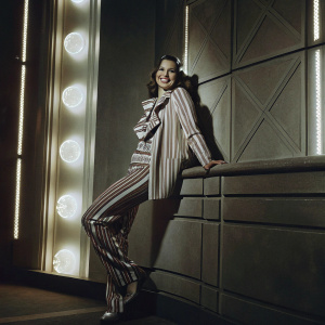 Hailee Steinfeld, radiant smile, elegant striped outfit, posing against neon-lit backdrop with futuristic designs. Studio portrait