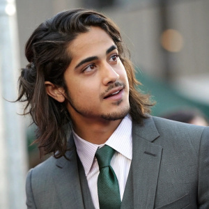 Avan Jogia, the actor, in a close-up. Gray suit and green tie. Long wavy hair. Slight stubble. Professional photograph with blurred background