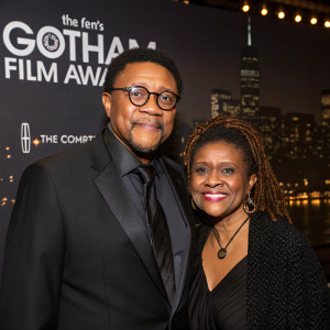 Greg Mathis and Linda Reese at the Gotham Film Awards. Greg Mathis, African American male, wears dark suit and glasses. Linda Reese, African American female, has a warm smile and braided hairstyle. They pose in front of a dimly lit backdrop with city skyline.