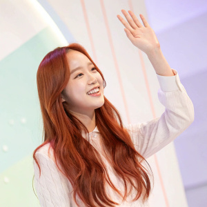 Kim Chaewon, young woman with long red hair, waving in a brightly lit studio. Soft pastel background with subtle gradient