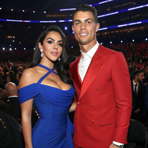 Cristiano Ronaldo and Georgina Rodriguez, dressed in elegant attire, attending the Grammy Awards ceremony. Georgina in a deep blue, off-the-shoulder gown. Cristiano in a sharp red blazer and white shirt. The venue is filled with celebrities and fans.