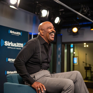 Steve Harvey in vibrant mood, laughing heartily in studio. Black shirt, gray pinstripe pants. SiriuxXM logos in the background. Studio lights
