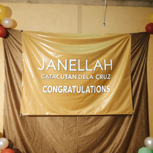 A vibrant congratulatory banner for Janellah Catacutan Dela Cruz, created by VLAG Designs. Festive backdrop with balloons and confetti.