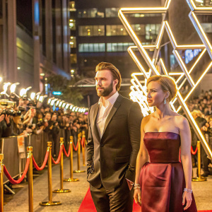 Chris Evans, sporting a sharp black suit and white shirt, stands beside Scarlett Johansson, elegantly dressed in a maroon strapless gown. They are at a crowded film festival with flashing cameras and vibrant red carpet