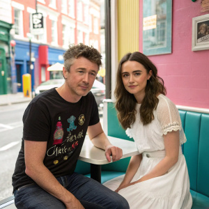 Aidan Gillen and Olivia O'Flanagan, Irish actors, in casual attire. Aidan in a black graphic tee, Olivia in a white dress. At a trendy cafe, surrounded by colorful walls and urban street scene.