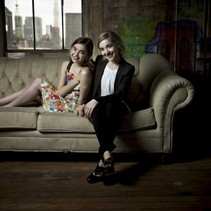 Rowan Blanchard and Kiernan Shipka posing for a candid photo shoot. The background is a vibrant cityscape with colorful graffiti walls. Rowan wears a bright floral dress, while Kiernan opts for a chic black blazer over a white blouse. Both are seated on a vintage green sofa in a cozy studio setting.